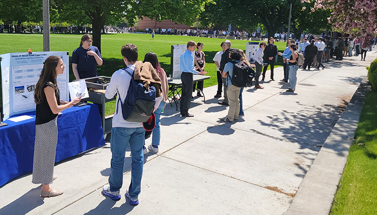 Sidewalk with multiple people at tables on a sunny day, looking at academic posters and talking Sidewalk with multiple people at tables on a sunny day, looking at academic posters and talking