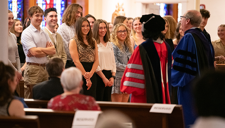 A group of students stands in front of people in graduation robes A group of students stands in front of people in graduation robes